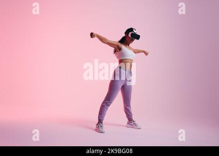 Woman exploring a virtual reality game in a studio. Active young woman standing with her arms outstretched while wearing virtual reality goggles. Fema Stock Photo