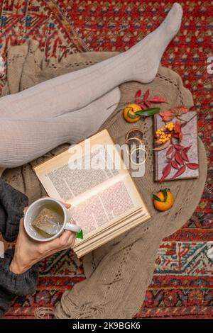 The legs of a woman sitting on a carpet with wool socks and a cup of herbal tea in her hand, while reading a book Stock Photo