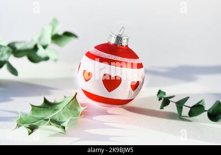Single Christmas decorative trinket. Red white glass ball with heart shapes. Fragrant holly and eucalyptus twigs. Sunlight, long shadows, off white Stock Photo