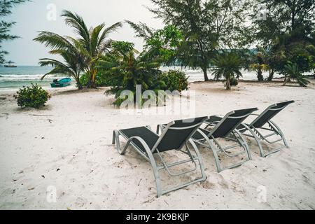 Tropical resort with chaise longs arranged in row under palms on sandy beach. three sitting chairs on the island among the palm trees under the hot su Stock Photo
