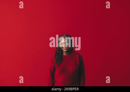 Black woman with dreadlocks laughing happily while standing against a red background. Cheerful mature woman embracing her natural hair with pride. Stock Photo