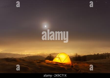 Yellow tent lighted from the inside against the backdrop of glowing city lights in fog. Amazing snowy landscape. Tourists camp in winter mountains. Travel concept Stock Photo