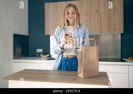 Young woman unpacking paper bag after shopping using smartphone in kitchen interior at home, copy space. Stock Photo