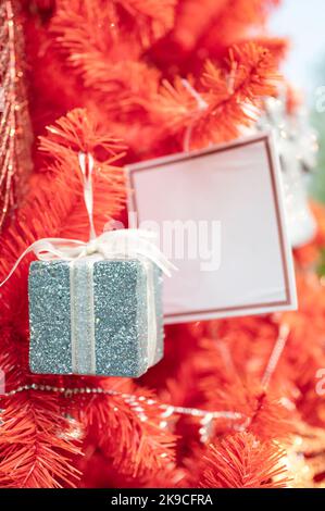 Gift box with greeting card hanging on red christmas tree Stock Photo