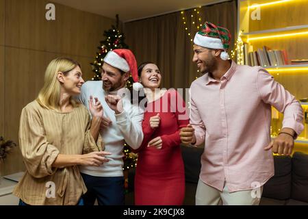 Four diverse friends at New Year's party celebrating and dancing, house guests having fun at Christmas, in New Year's festive clothes and Santa hats in the living room. Stock Photo