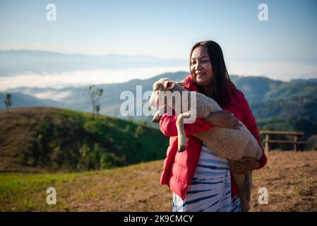 Young Asian woman is hugging intimately a little lamb at sheep farm in the morning with beautiful landscape. Stock Photo