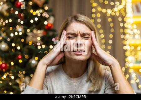 A young beautiful woman is sitting at home, holding her head with her hands, grimacing in pain. She feels a severe headache. She is tired of preparing for the New Year and Christmas holidays. Stock Photo