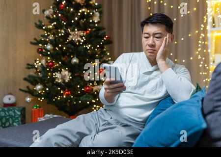 Worried asian man read bad news message notification on new year christmas eve day looking at mobile phone smartphone screen Disappointed despair male sitting at home background Christmas tree. Stock Photo