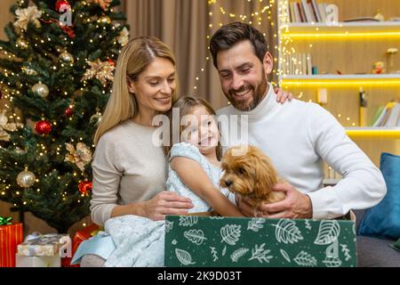 Happy parents make a surprise gift of a small dog puppy from Santa to a child for Christmas or New Year. Happiness. Father and mother surprising their kid little girl receives a present at home. Stock Photo