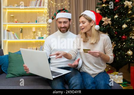 Happy family couple man and woman on Christmas do online shopping in online store sitting on sofa at home near Christmas tree, man holding bank credit card in hands, using laptop for remote shopping. Stock Photo