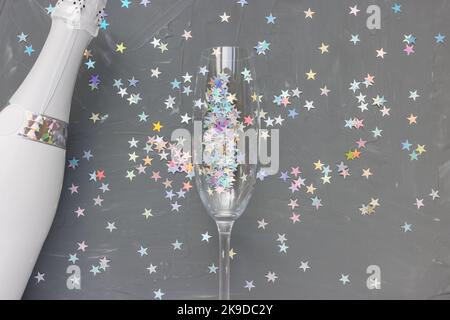 White Champagne bottle and flute glass on black with colorful stars confetti festive background. Winter holiday, New Year party and celebration concept. Creative flat layout with copy space Stock Photo