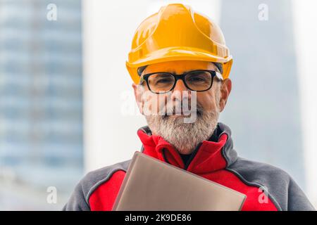 Portrait of a confident, caucasian, bearded, grey-haired, middle-aged professional construction worker in uniform wearing a bright orange helmet and eyeglasses, holding a folder with documents, looking at the camera. High quality photo Stock Photo