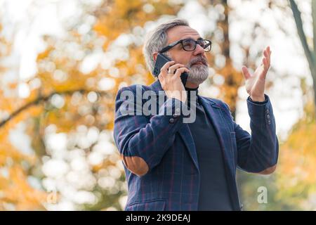 Middle-aged businessman with grey beard and hair wearing glasses and jacket talking expressively on the phone walking around park. Horizontal outdoor shot. High quality photo Stock Photo