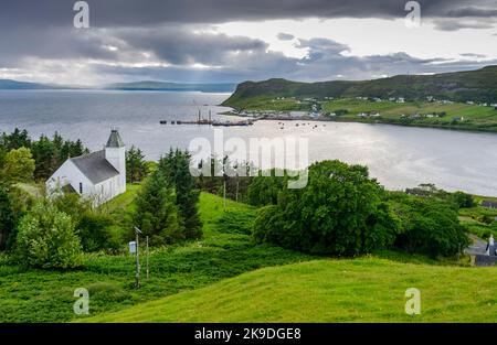 White painted,Ssottish Presbyterian church,on a green,grassy hill,surrounded by fir trees at edge of the sea,Uig harbour and ferry terminal across the Stock Photo