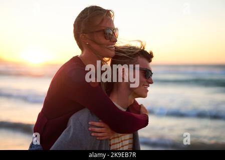 Being together makes them happy. an affectionate young man piggybacking his girlfriend on the beach. Stock Photo