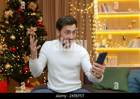 A happy young handsome man is sitting on a sofa in a New Year's decorated apartment. He holds the phone in his hands, rejoices, finds good pre-Christmas and New Year's discounts on gifts
