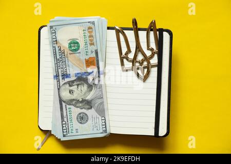 One hundred dollars lie in a white empty notebook on yellow and next to it lies the coat of arms of Ukraine, business and income in Ukraine, a notepad Stock Photo