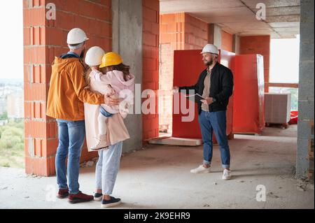 Man and woman with child standing inside apartment building under construction and talking with builder. Family future homeowners discussing building process with construction worker. Stock Photo