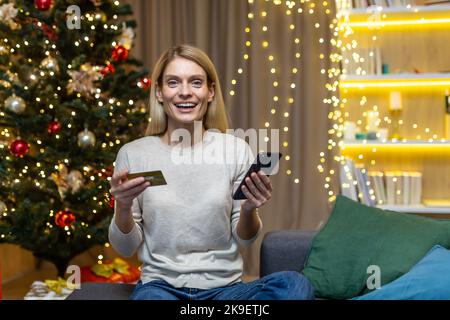 Portrait of happy online shopper woman, christmas woman looking at camera and smiling happy holding bank credit card and phone in hands, sitting on sofa near tree celebrating new year. Stock Photo