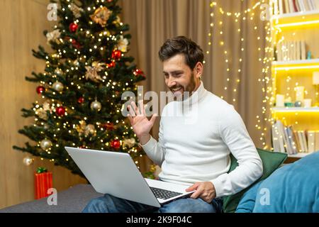 Man at home recording video greetings, Merry Christmas and Happy New Year, sitting on sofa at home near Christmas tree, using laptop for online remote communication with friends, video call. Stock Photo