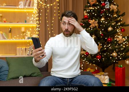Upset and disappointed christmas man reading online messages and notifications from smartphone, sitting at home on christmas sofa during new year holidays. Stock Photo