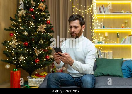 Serious and focused man using smartphone at home, businessman typing message near Christmas tree. Stock Photo