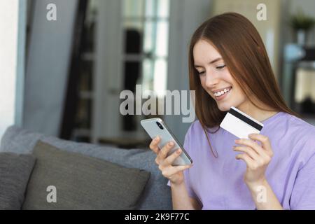 Happy smiling female holding credit card, looking at smartphone screen Stock Photo