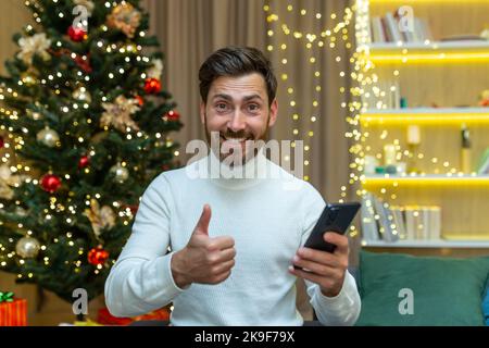 a man on Christmas looks at the camera and shows a finger up affirmatively holds a smartphone in his hands, sits at home on the sofa near the Christmas tree on New Year's holidays. Stock Photo