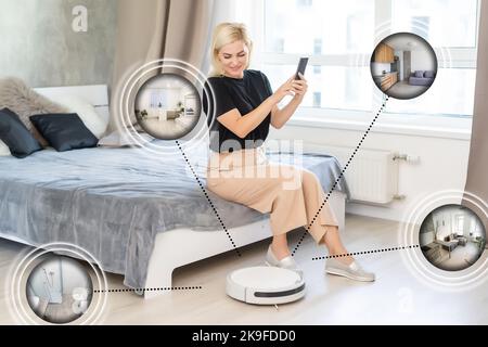 happy young woman using smart home application on smartphone and robot vacuum cleaning floor in background in the modern living room. Stock Photo