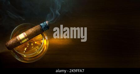 a glass of brandy on a wooden table, a steaming cuban cigar in a smoky atmosphere of a night bar Stock Photo