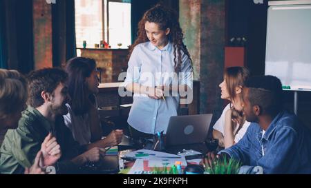 Young woman director of small company is talking to her employees holding business meeting at table in modern loft office. People, teamwork and management concept. Stock Photo