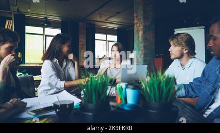 Young woman is speaking with colleagues sitting at table in modern office discussing work plans while her team is listening to her, male coworker is drinking tea. Stock Photo