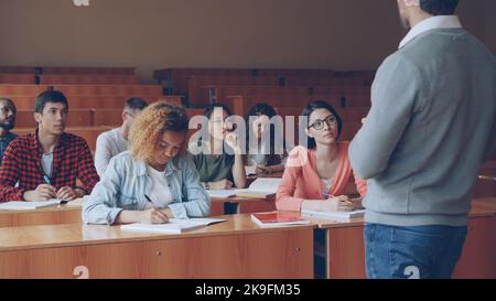 Emotional tutor is speaking to adult students explaining new information while attentive pupils are listening and taking notes. Profession, higher education and people concept. Stock Photo
