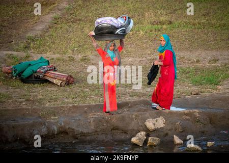 Two Egyptian women, one carrying metal pots on her head, dressed in bright red, washing pots, pans and carpets on the banks of the river Nile, Egypt Stock Photo