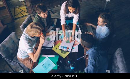 Multiracial workgroup is analyzing diagram and writing on colored stickers sitting at desk in office room. High angle view of people talking, pointing at charts and taking notes. Stock Photo