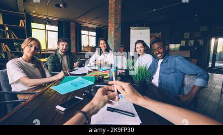 Office workers are smiling and laughing looking at camera during briefing with CEO sitting at desk in modern office during working day. Friendly atmosphere and teamwork concept. Stock Photo