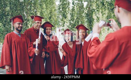 Cheerful guy is using smartphone to take pictures of his fellow students graduates holding diplomas, posing and laughing. Happiness, modern technology and education concept. Stock Photo