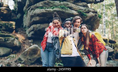 Multiracial group of friends modern tourists are taking selfie in forest with rocks in background using smartphone, men and women are posing and showing hand gestures. Stock Photo