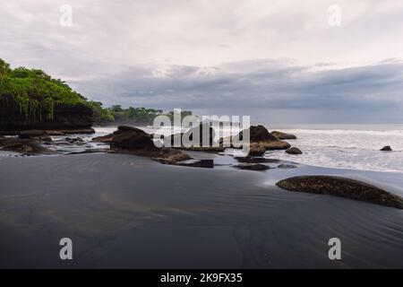 Volcanic sandy beach with ocean and waves in Balian, Bali. Beach with rocks and cloudy sky Stock Photo