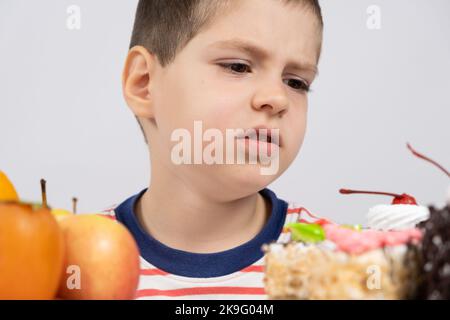 5 year old cute boy sits in front of fruits and cakes and chooses what to eat Stock Photo
