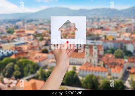 Woman's hand is holding a paper with a house cut out on it, panorama of the city in background. The concept of renting or buying real estate Stock Photo