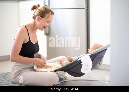 Happy smiling mother playing with her little baby son sitting in electric rocking chair at home. Child development and happy parenting. Stock Photo