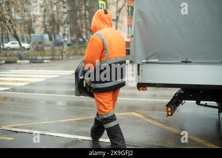 Worker puts cargo in transport. Orange clothes for work. Cleaning road. Man throws bag. Stock Photo