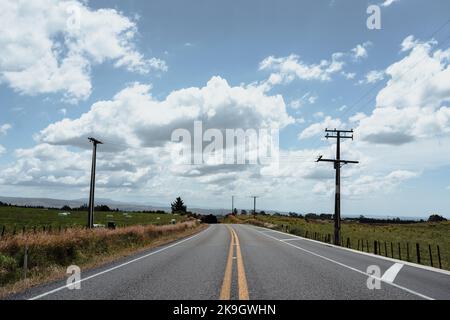 long asphalt road with no traffic next to electricity poles running through green meadows with lots of grass under a cloudy sky, rotorua, new zealand Stock Photo