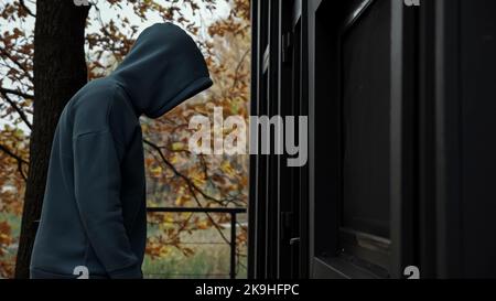 Unrecognizable man in dark clothes with hood picks door lock and enters house on evening Stock Photo