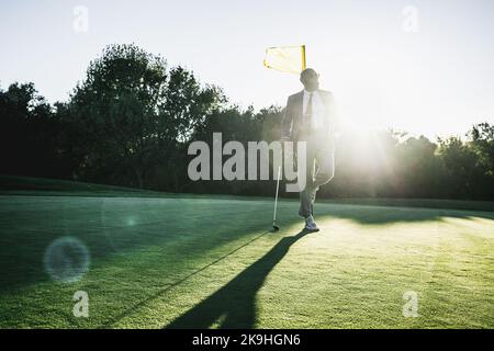 A dapper bald bearded black man in an elegantly tailored suit with a necktie and sunglasses is leaning against his club and a flagpole while standing Stock Photo
