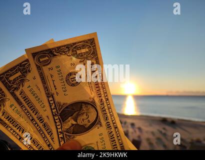 A man holding three one-dollar banknotes in his hand against backdrop of the sea, sandy beach and blue sky on sunny summer day. The concept of money, finance, monetary, travel, tourism, vacation, rest Stock Photo
