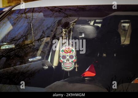 Hanging Day of the Dead cardboard sugar skull hangs with beads from rearview mirror of car with dark interior blurred in background- spooky tree limbs Stock Photo