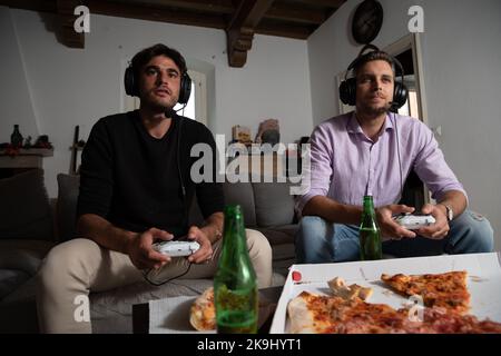 Concentrated friends playing hard and fast at video games with pizza and beer on the table. Stock Photo