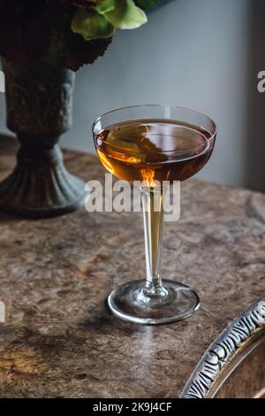 whiskey cocktail in hollow stem coupe glass on burl wood table with bouquet in a vase Stock Photo
